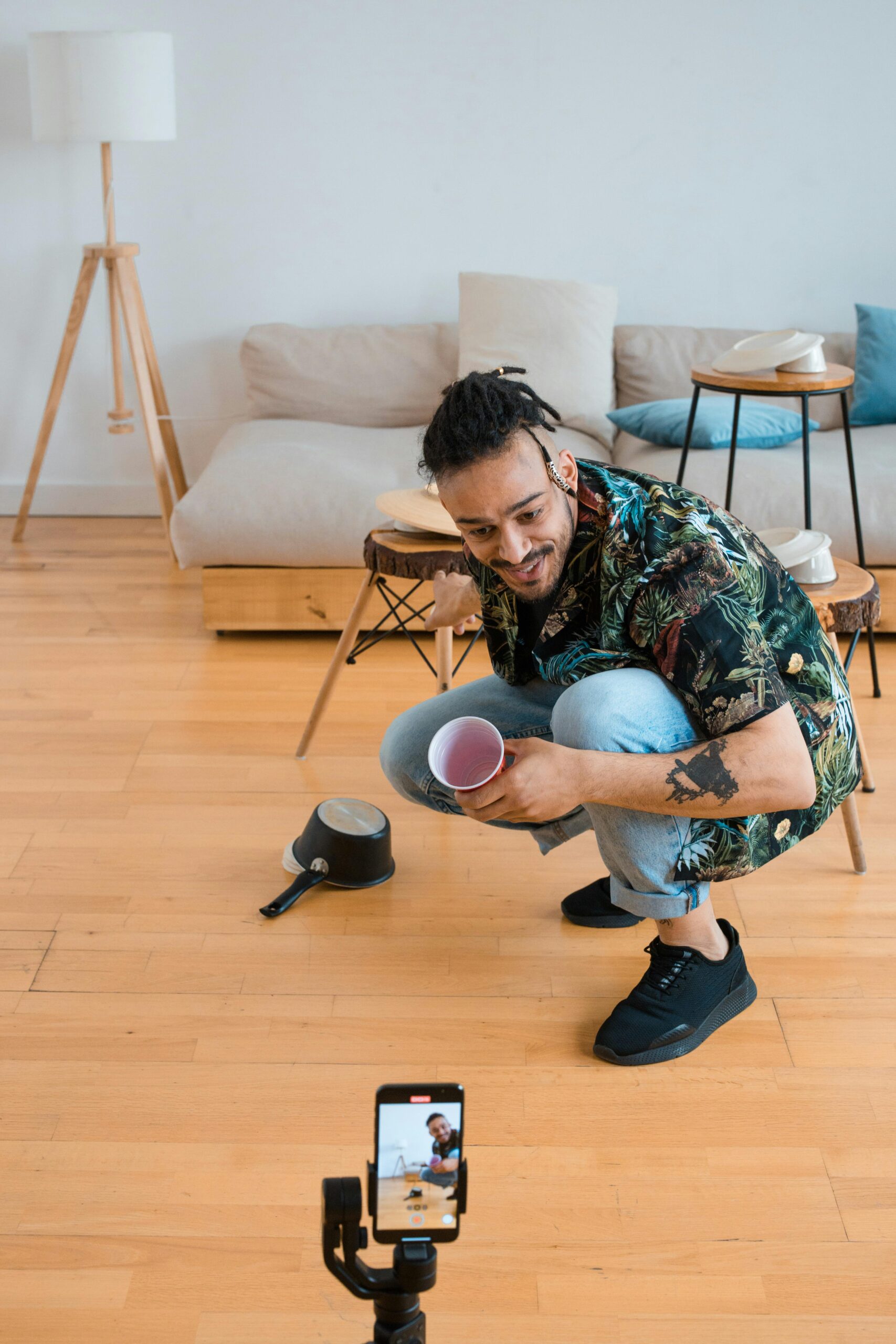 A young man squatting indoors, filming content using a smartphone in a modern living room.